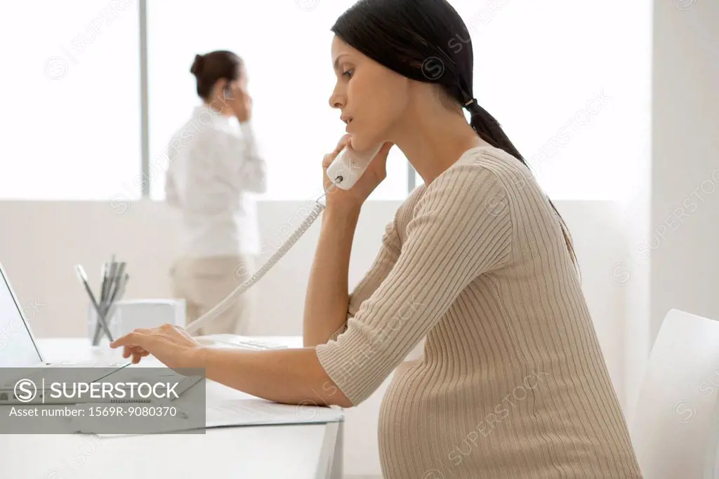 Young pregnant woman talking on landline phone in office, backlit