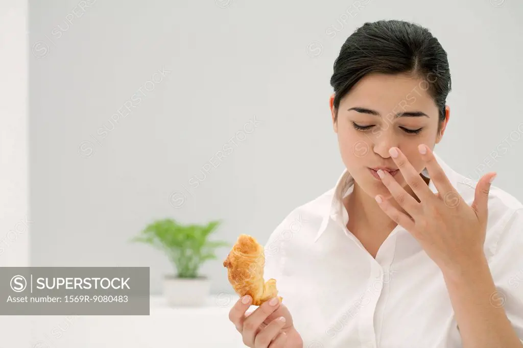 Young woman licking finger while eating croissant