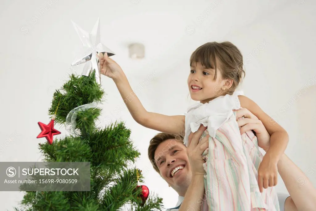 Father carrying little girl putting star on tip of Christmas tree