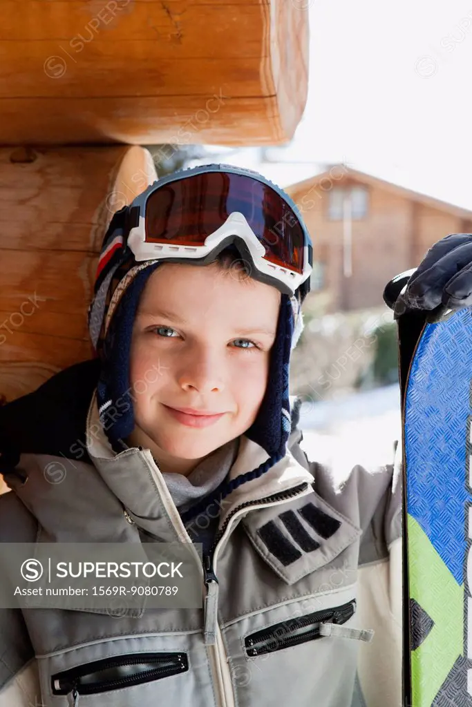 Boy wearing ski goggles and ski jacket and holding snowboard, portrait