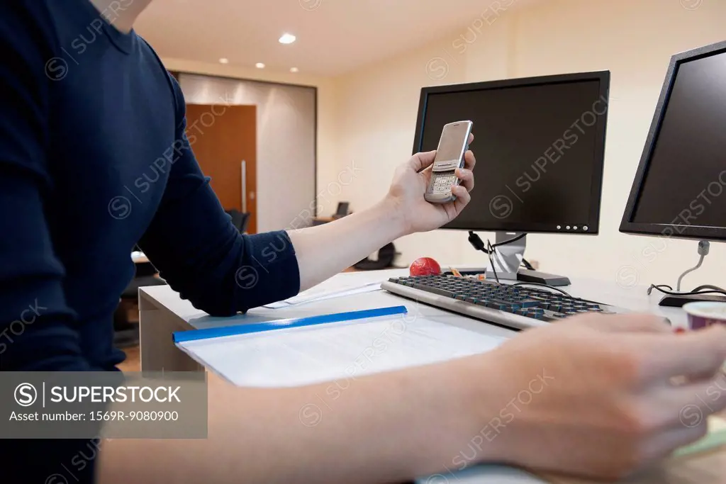 Man working at desk in office, holding cell phone, mid section
