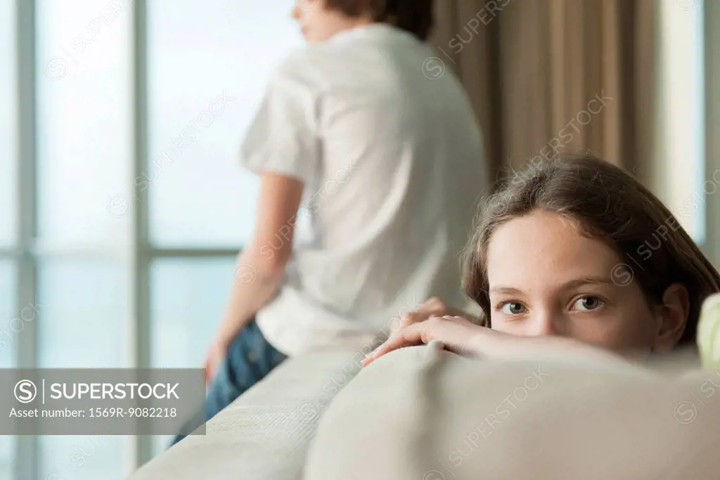 Girl sitting on sofa with head resting on arm, brother looking out window in background