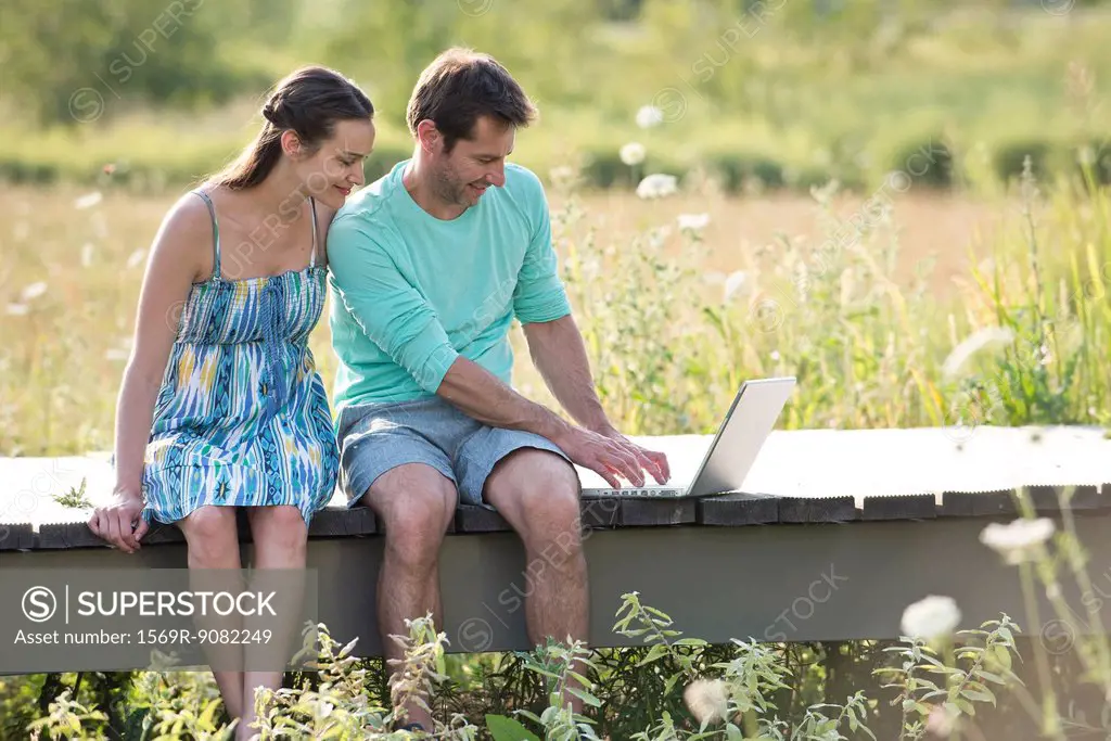 Couple sitting on countryside boardwalk with laptop computer