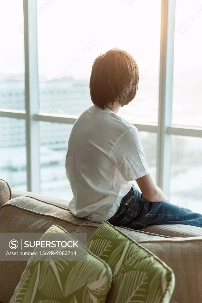 Teenage boy sitting on edge of sofa, looking out window, rear view