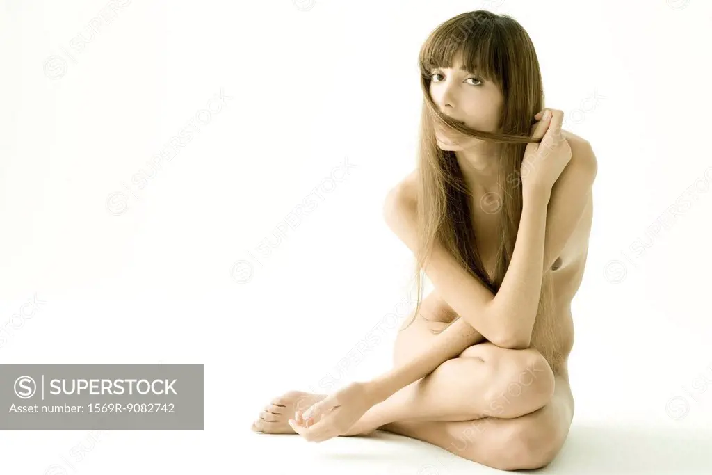 Nude woman sitting with arms folded across chest, covering mouth with strand of hair