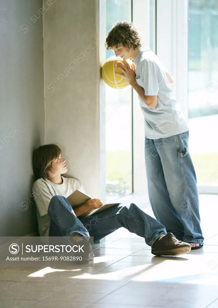 Boy sitting on floor studying, friend holding basketball, talking to him