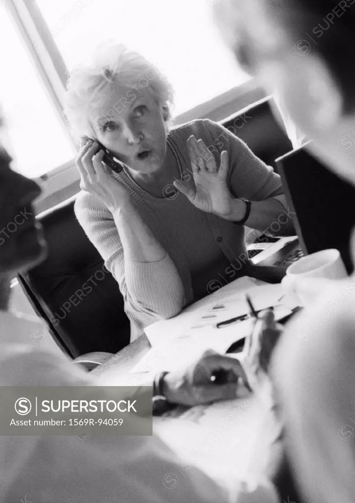 Mature businesswoman using cell phone, colleagues in blurred foreground, B&W