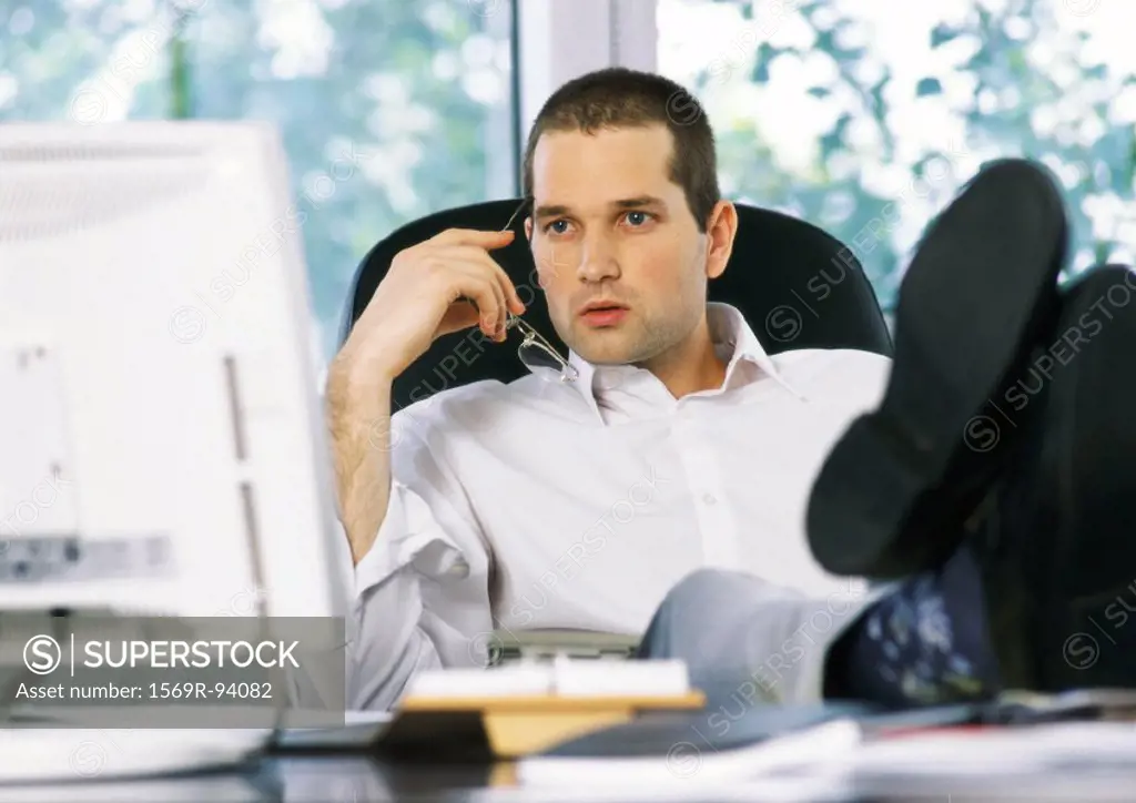 Businessman holding glasses next to face, looking at computer screen, sitting with feet on desk
