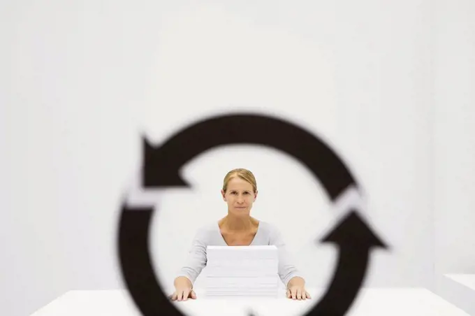 Professional woman sitting behind stack of paper, looking at camera through recycling symbol