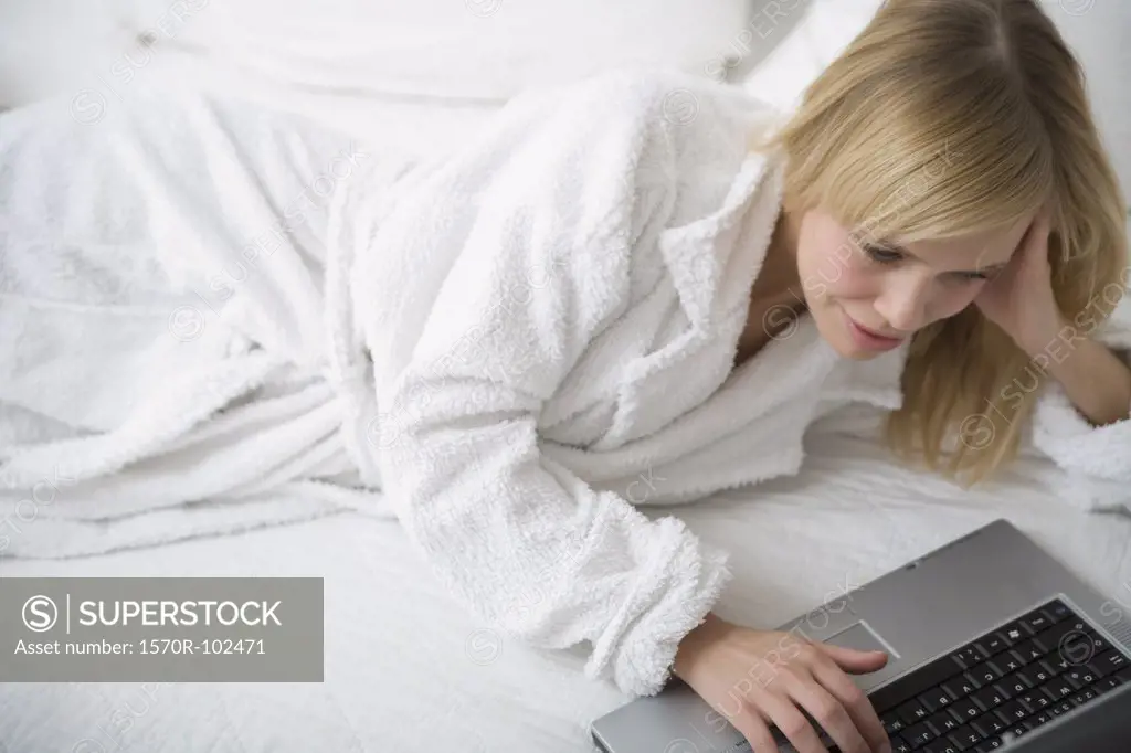 Young woman lying down on a bed in a bathrobe and using a laptop