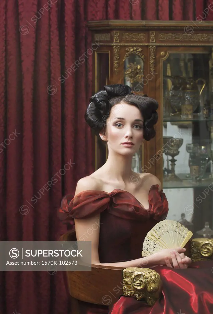 Elegant young woman seated in red ball gown and holding fan