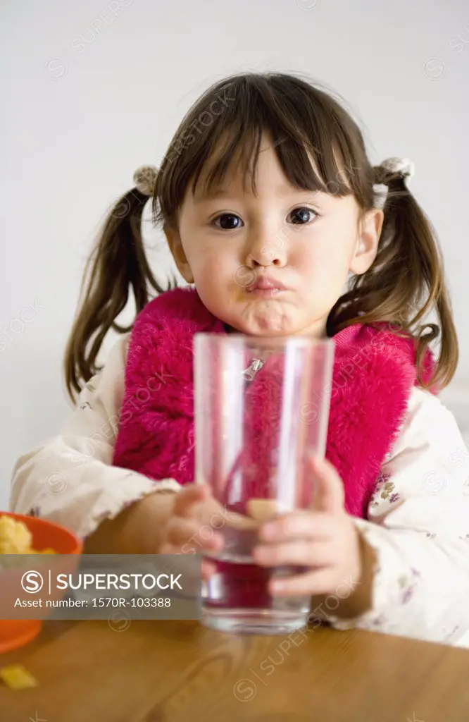 Young girl with pigtails sitting at table and holding glass of water