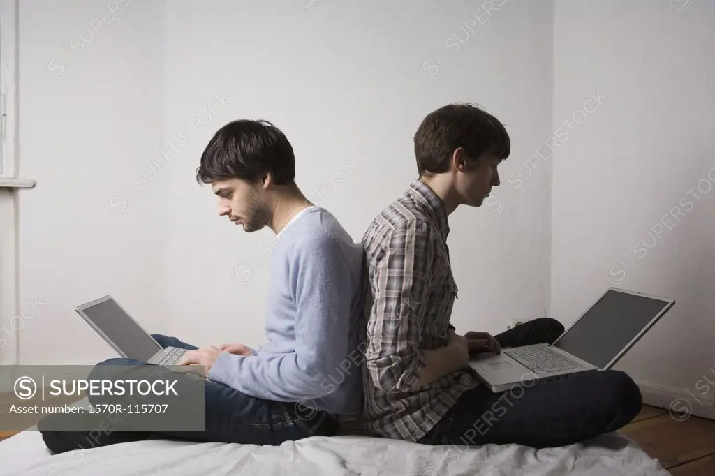 Two young men sitting back to back using laptops