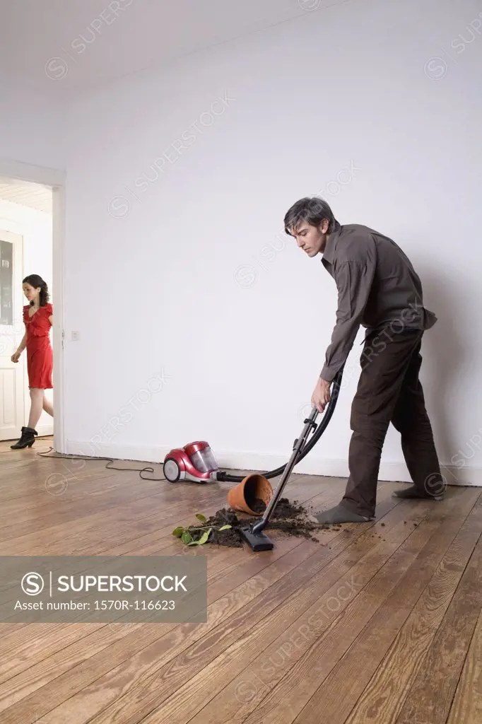 A man vacuuming up soil from a spilt potted plant whilst a woman is walking down a hallway