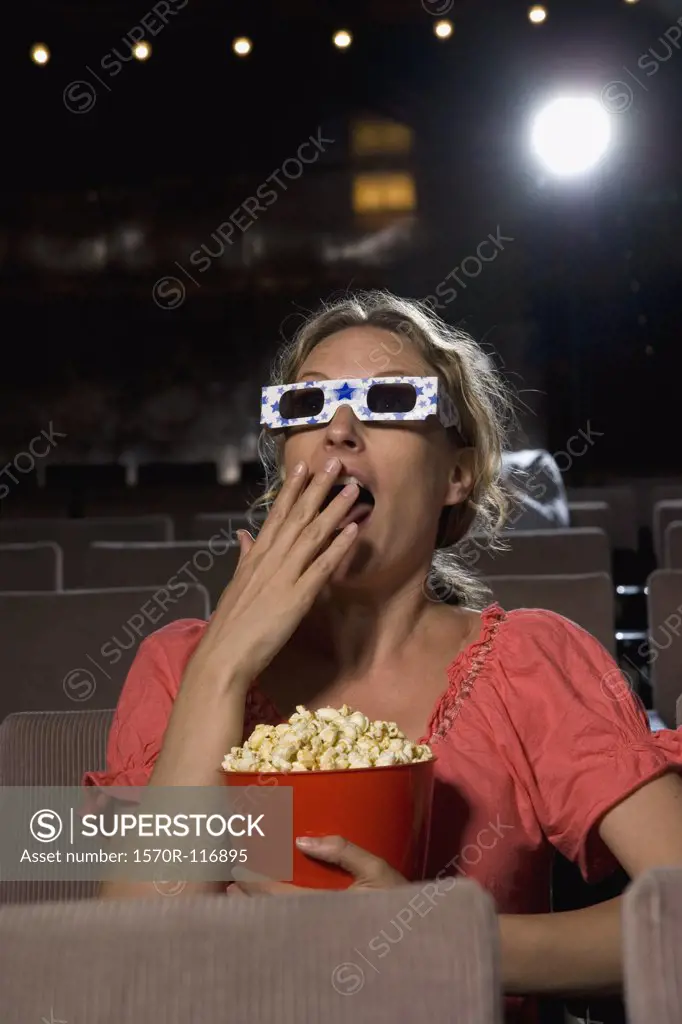A woman sitting in a movie theater wearing 3-D glasses and eating popcorn