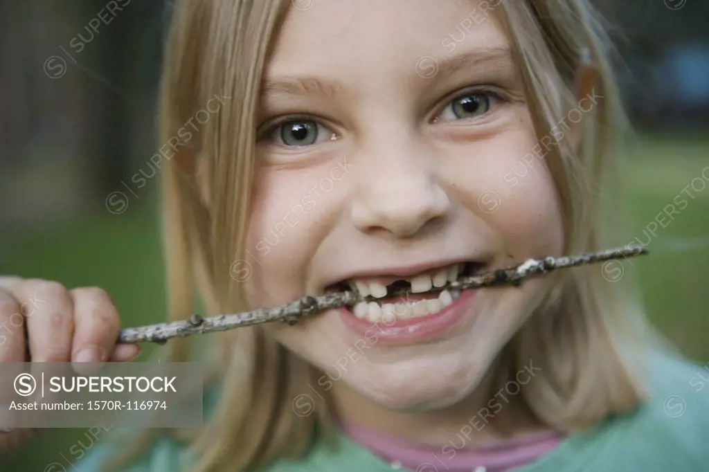 A young girl with a missing front tooth biting a stick