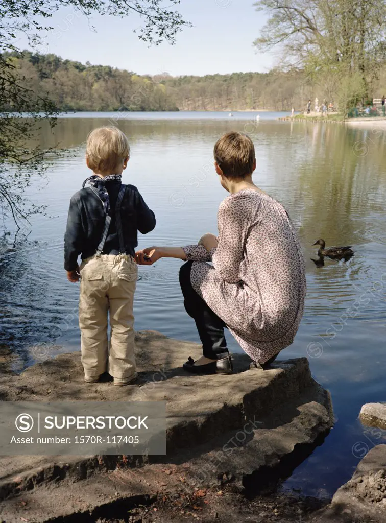 A mother and son feeding ducks by a lake