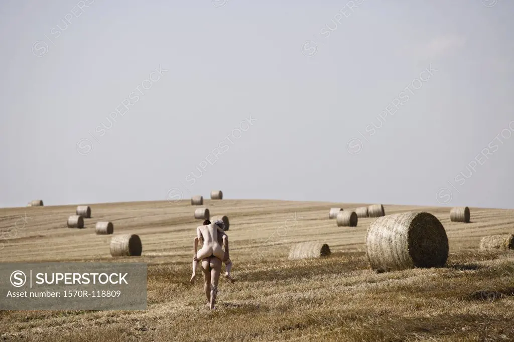 A naked man giving a naked woman a piggyback through a field