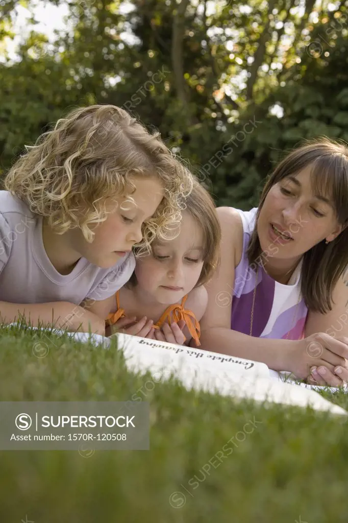 A woman and two girls lying down in a backyard and reading a book together