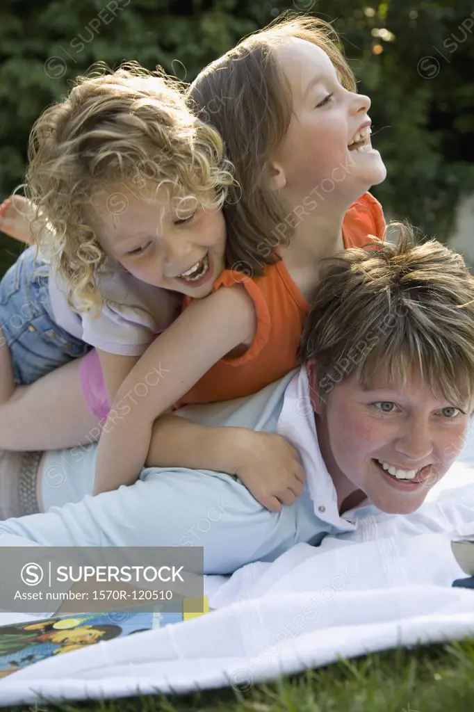 Two girls piled on top of a woman lying on a picnic blanket