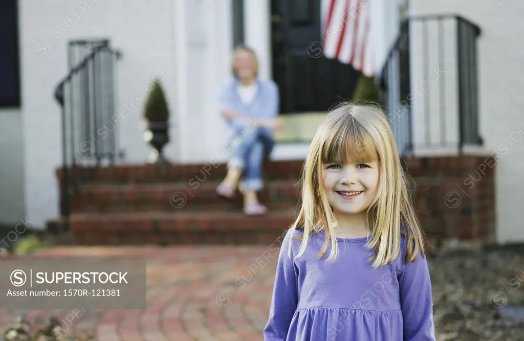 Young girl standing on a pathway in front of a house with her mother sitting behind on the doorstep