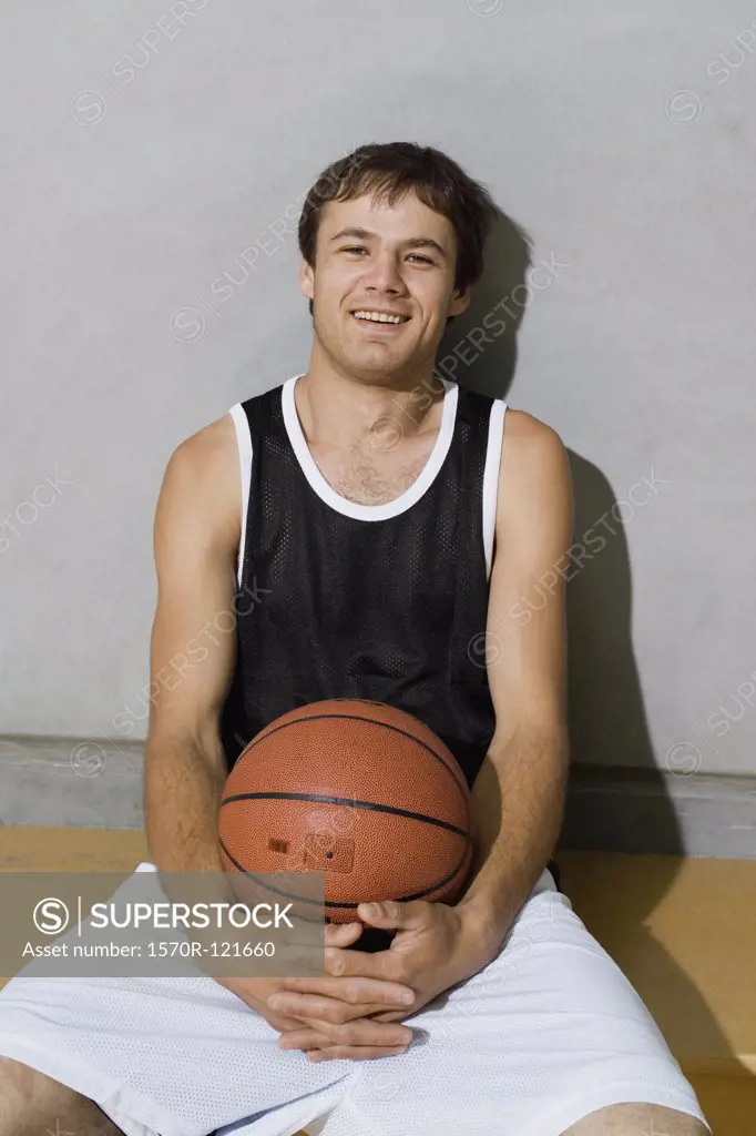 A young man sitting on a bench with arms around a basketball