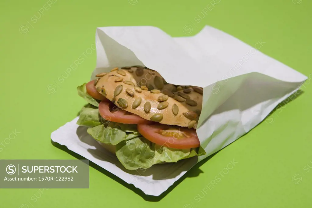A Sunflower seed bagel sandwich with tomato and lettuce in a paper bag