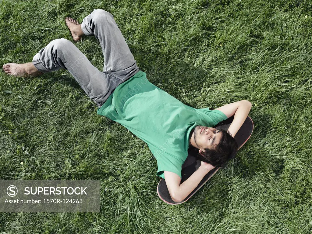 A young man lying in the grass with his head on a skateboard
