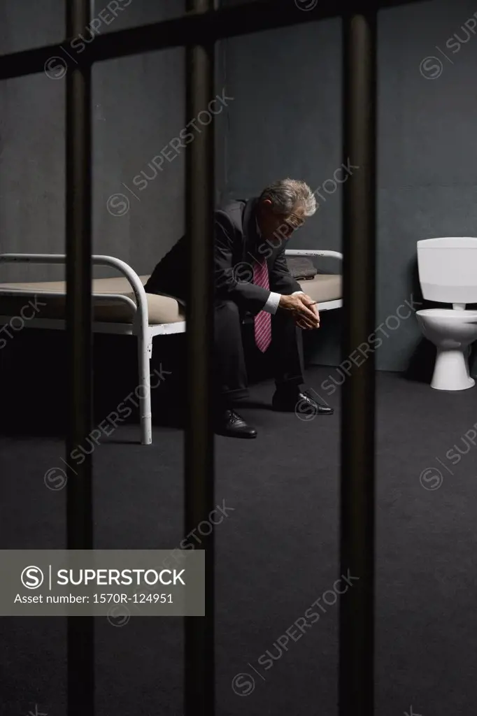 A businessman sitting on a bed in a prison cell