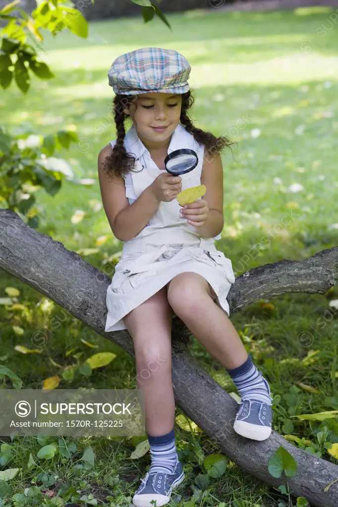 A girl sitting on a tree branch and looking at a leaf with a magnifying glass