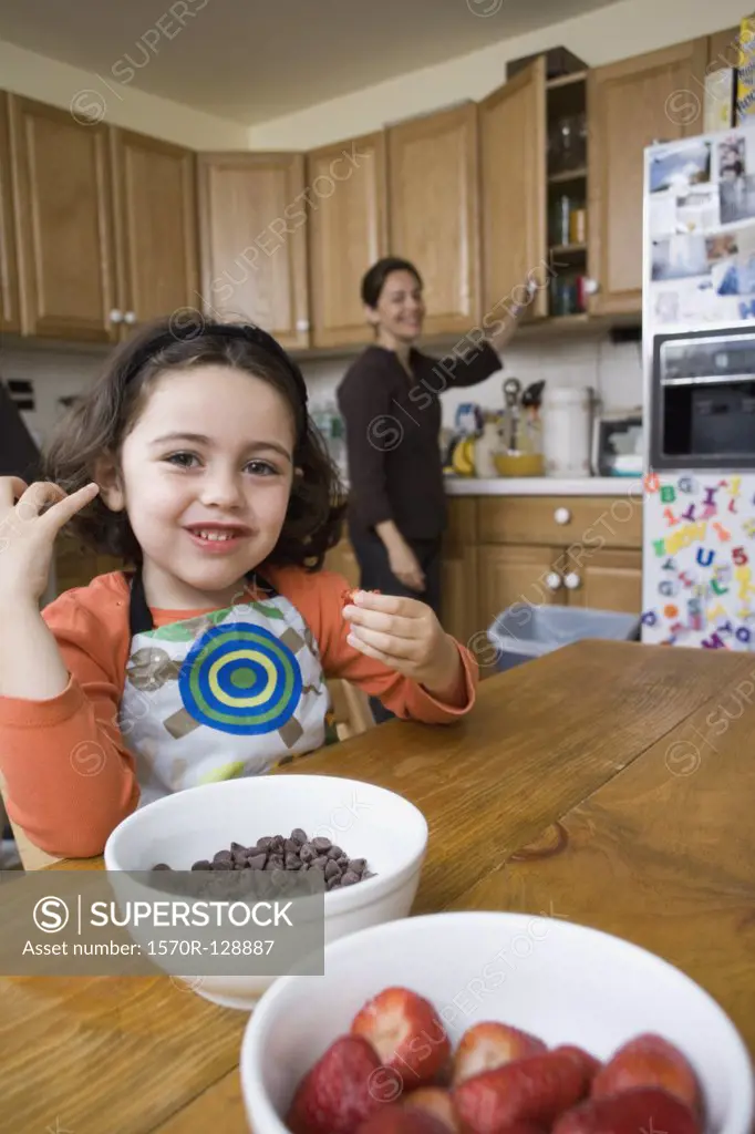 A young girl sitting at a kitchen table and her mother standing behind