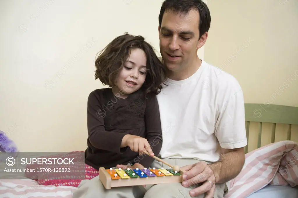 A young girl sitting with her father and playing the xylophone