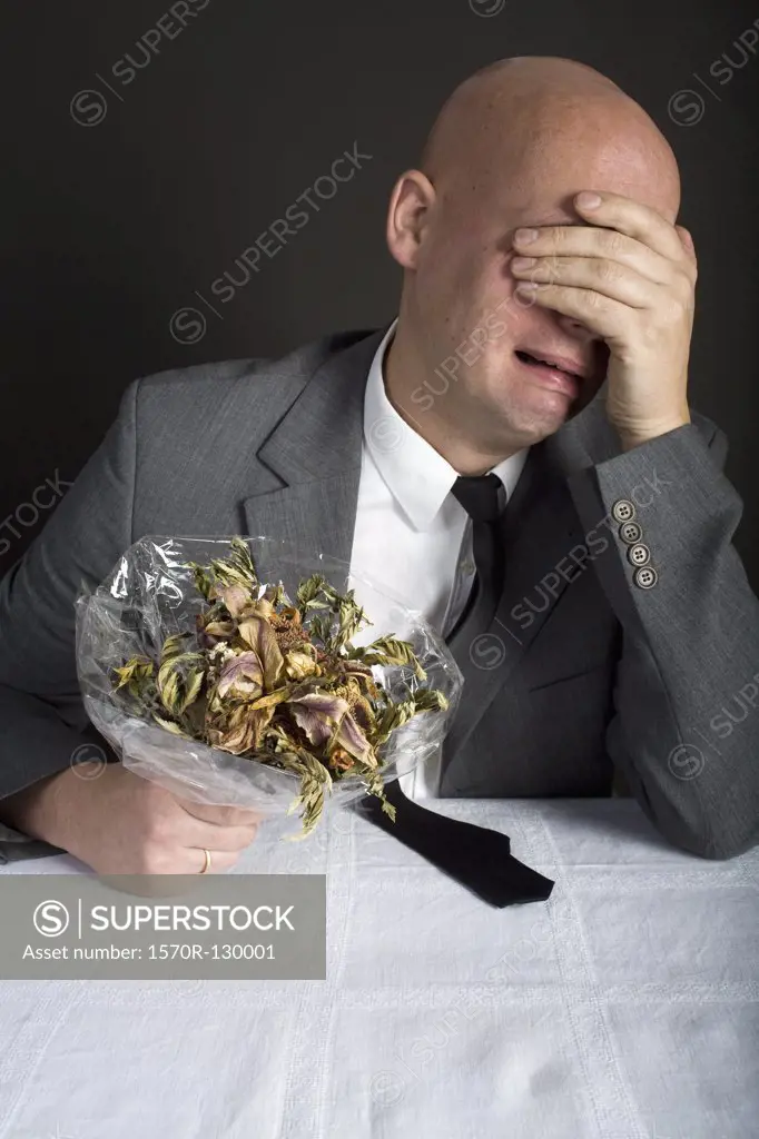 A well-dressed man holding a bouquet of dead flowers and crying