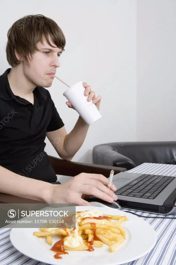 Young man using laptop whilst drinking soda and smoking a cigarette near plate of french fries