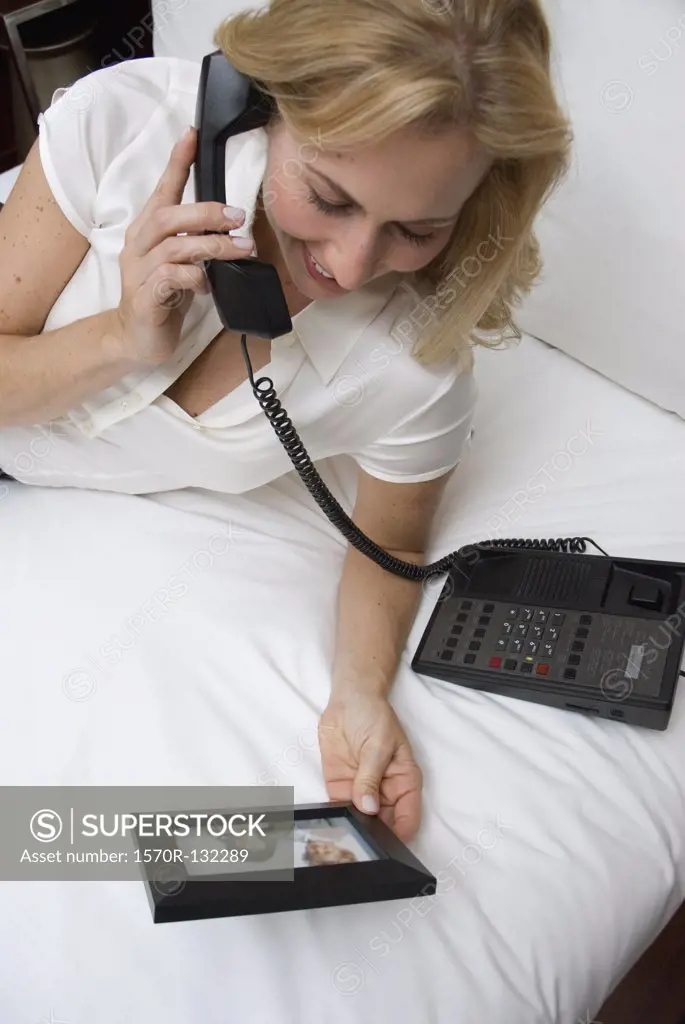 Woman looking at photograph of children whilst lying on bed in hotel room using telephone