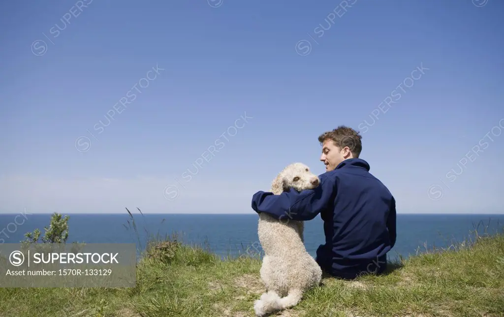 A man with his arm around a dog sitting by the sea