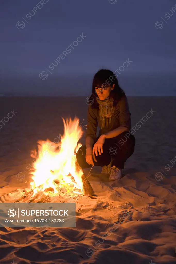 A woman staring into a fire burning on a beach