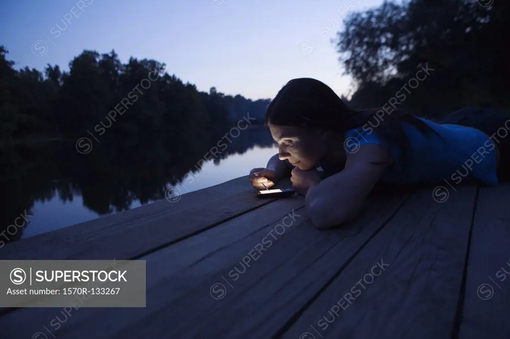 A woman using an electronic organizer while lying on a jetty, dusk