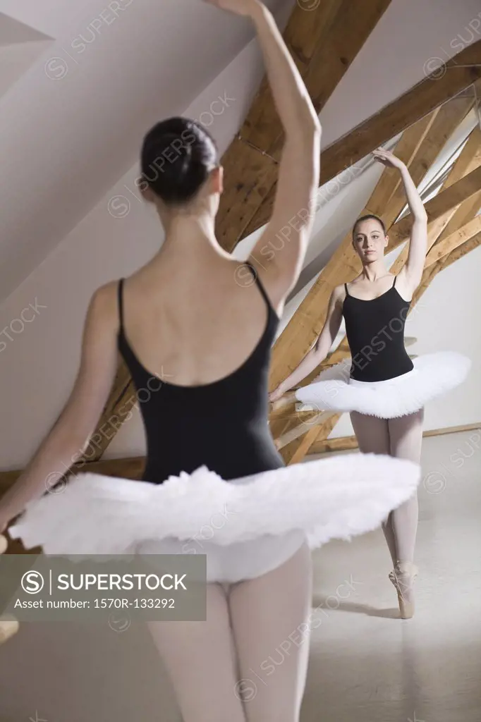 A ballet dancer on pointe with one arm raised in front of a mirror in a ballet studio