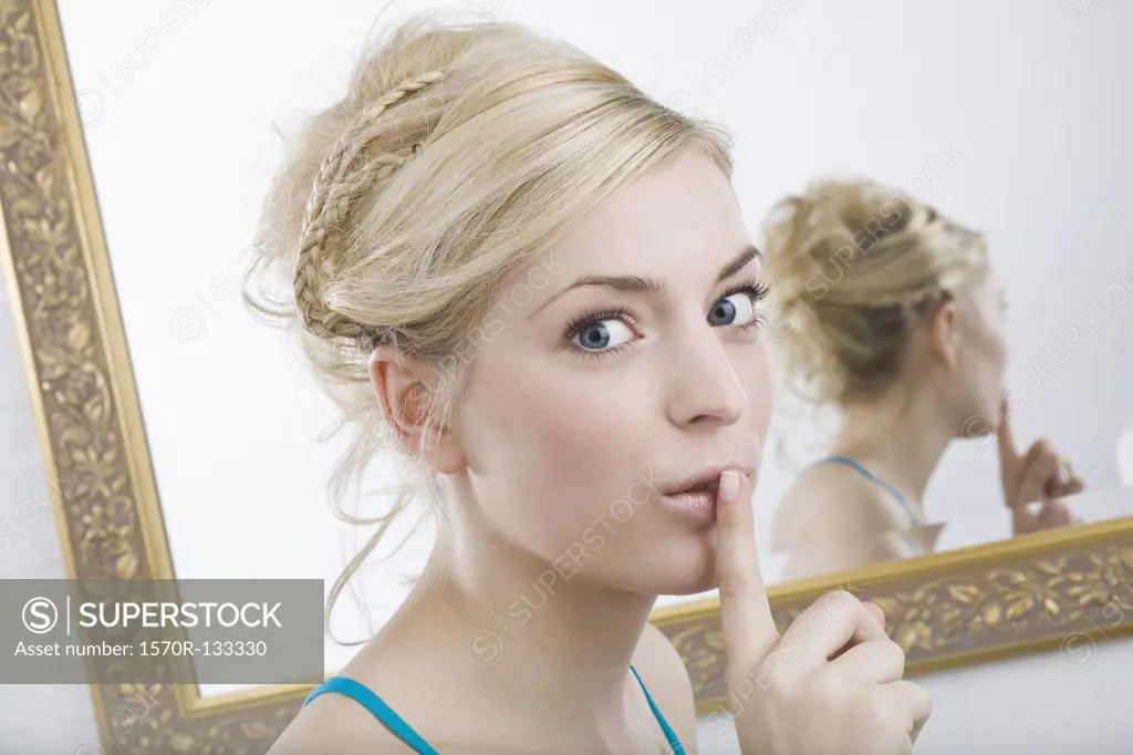 A woman with her finger to her lips in front of a mirror, looking at camera
