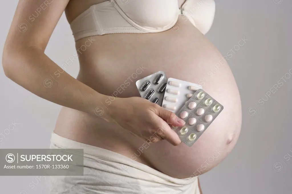 A pregnant woman holding blister packs of vitamins and nutritional supplements, midsection