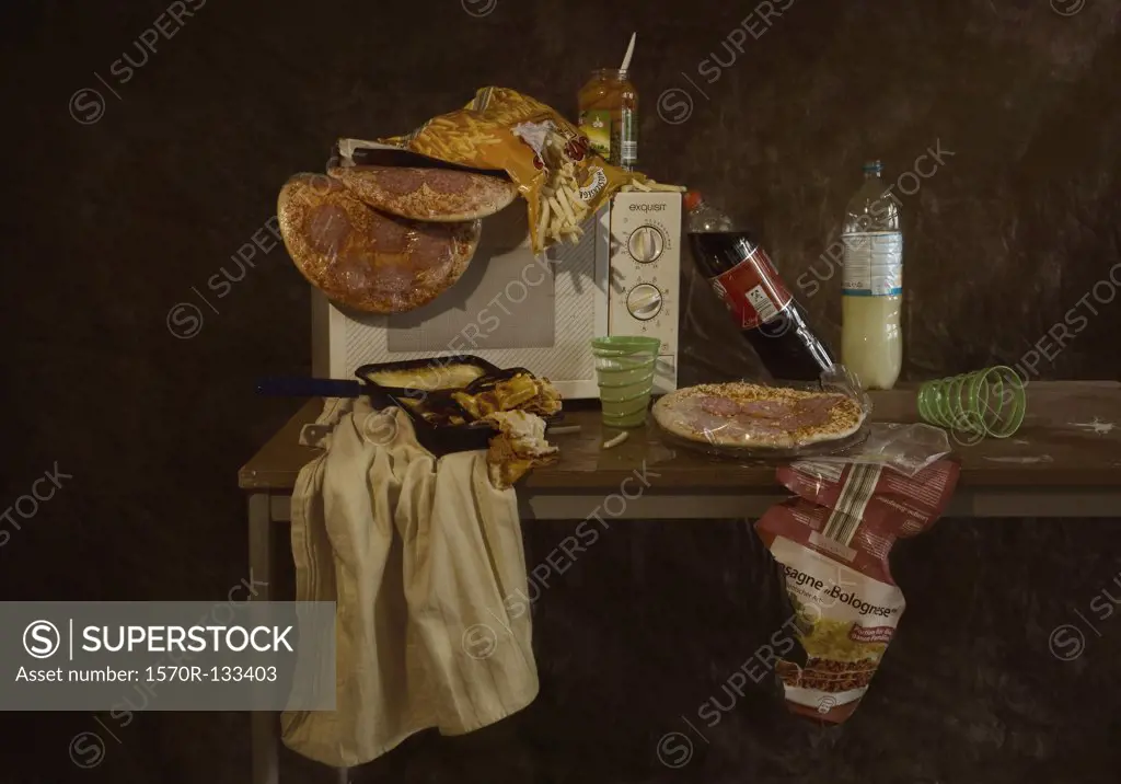 A table with a microwave and various frozen pizzas and frozen lasagna, still life