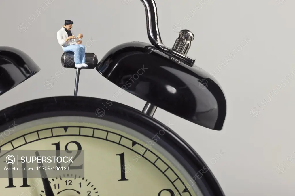 A miniature father figurine holding his baby while sitting on an alarm clock