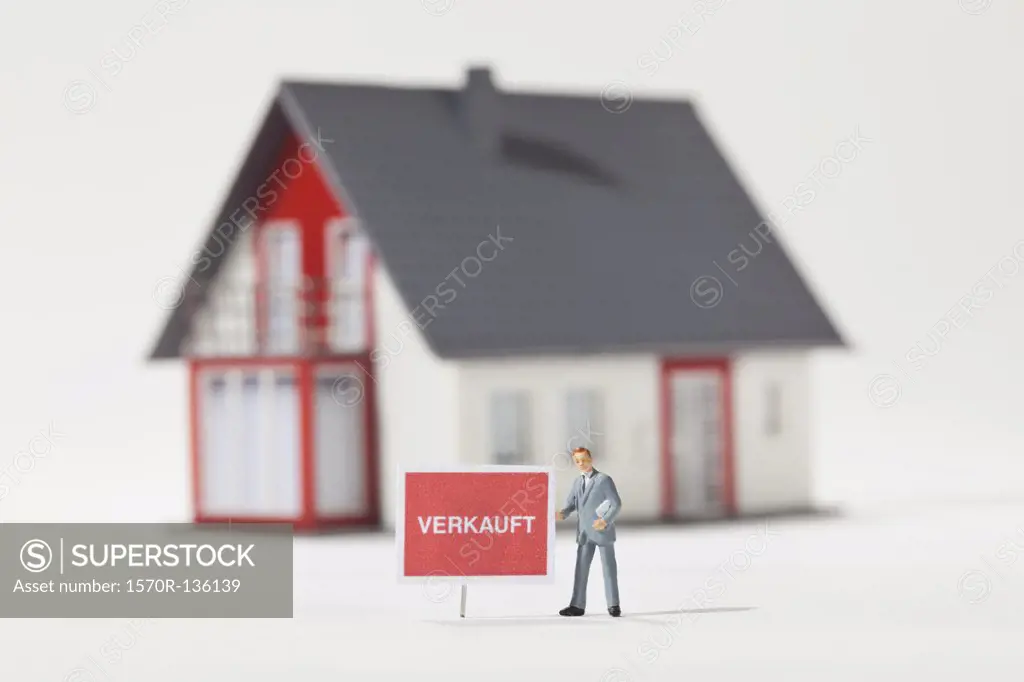 A miniature real estate agent figurine standing next to a VERKAUFT (sold in German) sign