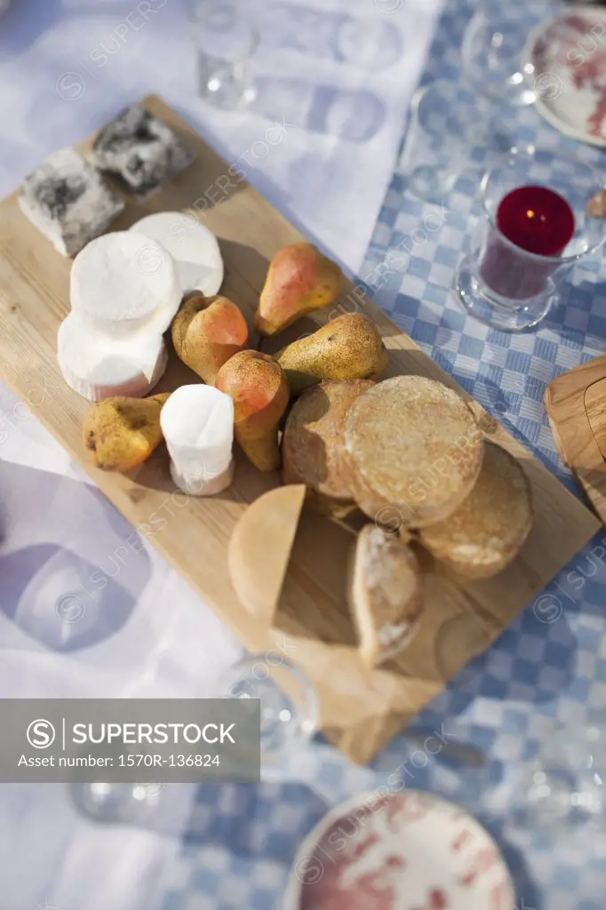 Variety of cheeses on a chopping board sitting on a table outside among glasses.