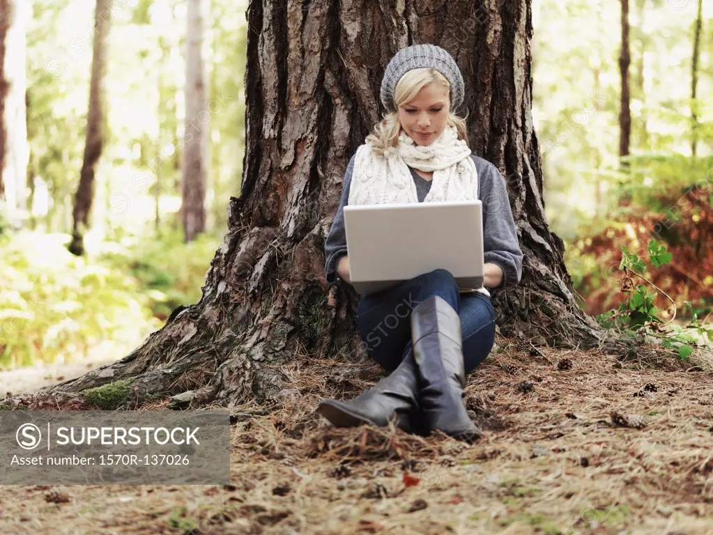 A woman in nature leaning against a tree and using a laptop