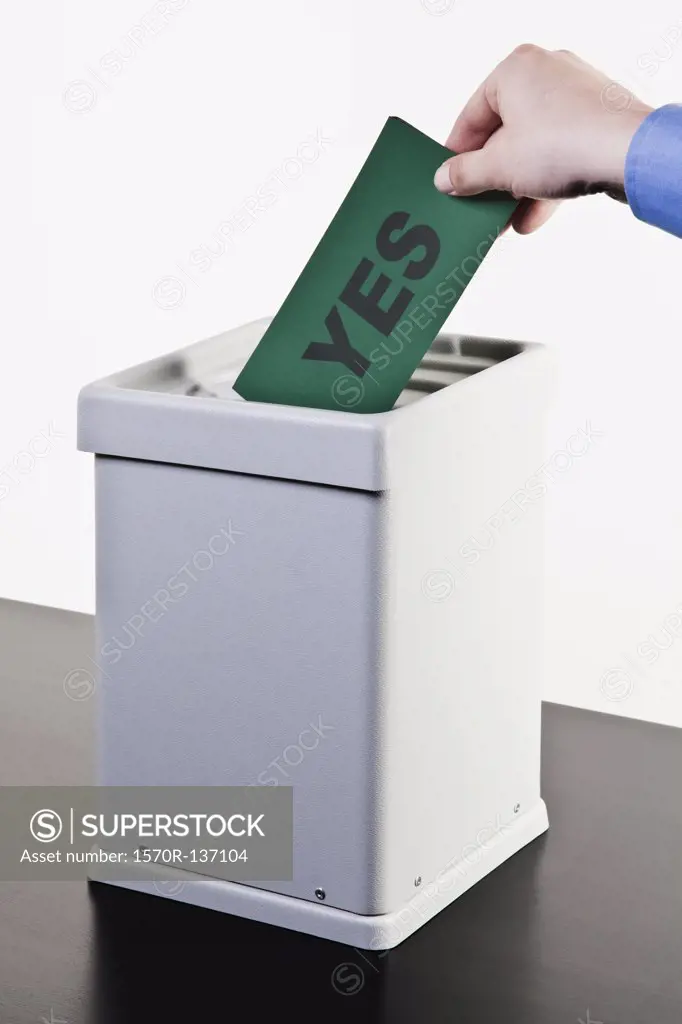 A man putting a ballot with the word YES written on it into a ballot box, close-up hands