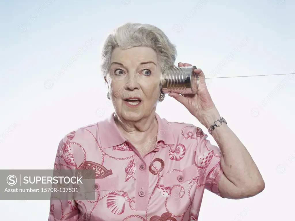 Senior woman comically strains to listen as she holds a tin can phone to her head