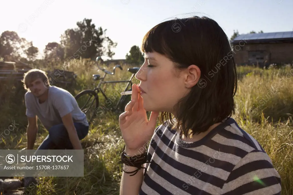 Girl smoking in field as guy in background watches