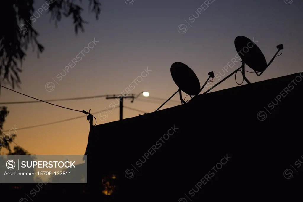 Silhouetted satellite dishes on a rooftop, dusk
